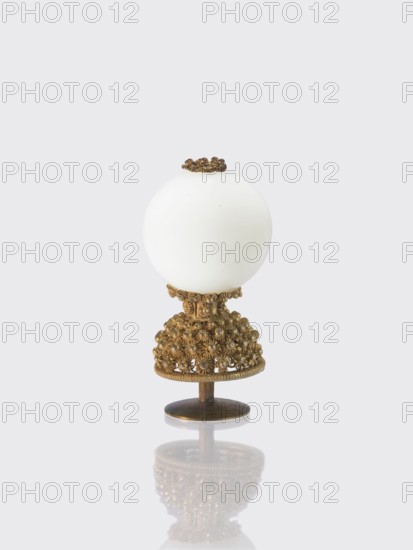 Button for a semi-official cap of a sixth-ranking official., Chinese art; Chinese culture. Qing dynasty 1644–1911. Officials; emblems. Button composed of a milky-white glass ball mounted on a pin with a dome decorated with rosettes. 19th century, China. Clothing, jewelry. Height 6.4 cm; diameter 3.2 cm., A decorative object composed of a smooth white spherical top mounted on an ornate metallic stand, with the stand featuring an intricate cluster of small rounded decorative elements and filigree details in a warm golden-bronze color, a small circular base in a darker bronze supporting the piece, and a subtle reflection of the object visible on the glossy surface beneath; visible colors include white, golden-bronze, darker bronze, and pale gray in the background.