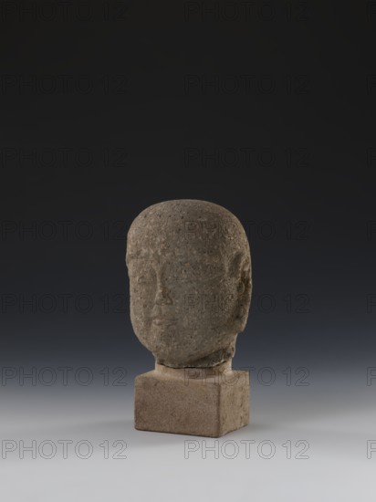 Head of a monk Buddha?, unknown sculptor, buddha iconography, Chinese art, Chinese culture, Ming 1368–1644, Chinese Buddhism, arhat/Luohan iconography, 14th–15th century, China, sculpture; height 11.8 cm, width 9.2 cm, depth 8.7 cm., A stone sculpture of a human head with a rounded top sits on a rectangular stone base, both surfaces showing a rough, pitted texture; the head appears in shades of gray and brown, and the base is a lighter brownish-gray. The background transitions from very dark gray at the top to a medium gray toward the bottom, and the ground surface beneath the base is a pale gray. The facial features on the head are subtly carved and worn, giving muted contours for eyes, nose, and mouth, and the overall scene includes tones of gray, brown, and subtle earthy speckles on the stone.