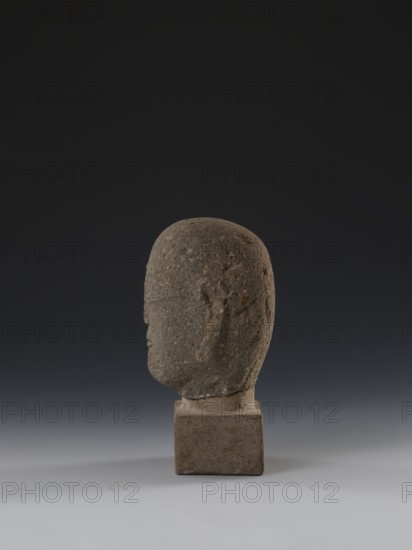 Head of a monk Buddha?, buddha iconography, Chinese art, Chinese culture, Ming 1368–1644, Chinese Buddhism, arhat iconography, Luohan iconography, 14th–15th century, China, sculpture, height 11.8 cm, width 9.2 cm, depth 8.7 cm, A weathered stone head sculpture in profile sits on a square stone base against a dark gradient background. The sculpture is textured with a rough surface showing small pits and irregularities, and the overall color palette includes shades of gray, brown, and muted beige on the stone, with the base a slightly warmer brownish-gray. The background transitions from nearly black at the top to a softer gray near the bottom, and the surface beneath the base is a light gray. The head has subtle carved facial contours visible from the side, including a rounded cranium, a brow line, a nose, and a hint of lips, with shadowing that gives a sense of depth.