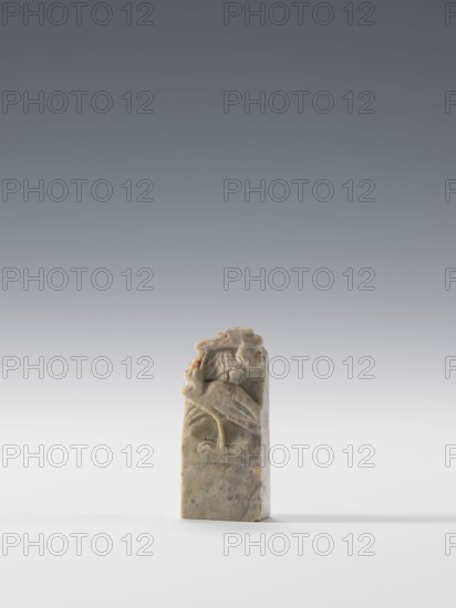 Seal stamp, Chinese, Qing 1644–1911, 19th/20th century. Blind stamp with a rectangular cross-section; bas-relief and engraved decoration depicting cranes. Seal stamp; height 6.5 cm, width 2.85 cm, depth 1.2 cm., A small rectangular carved object stands upright on a smooth surface, showing a relief of a stylized bird with a curved beak and wings folded against its body; the carving includes rounded, organic shapes and shallow grooves. The object appears to be made of stone with a mottled appearance in shades of light gray, pale green, beige, and small areas of brown, with subtle variations and speckling throughout. The background is a smooth gradient from darker gray at the top to lighter gray near the middle and almost white near the bottom, and a soft shadow falls to one side of the base.
