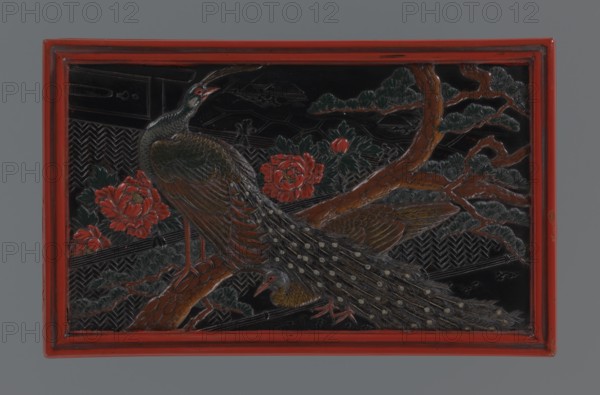Box with a lid, Japanese art, Japanese culture, Meiji 1868–1912, boxes, lacquer, peacocks, pines, peonies. Decoration on the lid: two peacocks in a chrysanthemum garden, pine tree, landscape in the background. Red, black, and dark green lacquer. 19th/20th century Meiji period, Japan. Container. Height 8.9 cm, width 20.2 cm, depth 12.5 cm, weight 369 g., A rectangular carved panel with a red outer frame shows a scene of two birds perched on a branch amid foliage and flowers; the birds have patterned plumage with dark greenish, brown, and gray tones and pale spots on their long tails, and small red accents near their heads and legs; the branch is brown with textured bark and supports green leaves and pine-like needles, while a few large flowers in shades of red bloom nearby against a predominantly black background with carved geometric patterns.
