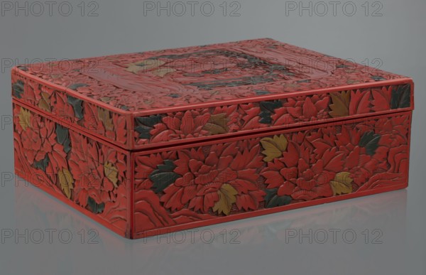 Box with a lid, Chinese art, Chinese culture, figural scenes, floral motifs, Qing 1644–1911, peonies, bamboos, geometric ornament, gourds, sages, scholars, fans, men. Decoration: on the lid a large reserve with a figural scene in a landscape; the rest of the background and sides—floral motifs. Red, black, green, and yellow lacquer, carved. 19th century, China. Container; height 13.0 cm, width 36.5 cm, depth 28.5 cm., A rectangular decorative box sits on a reflective surface, carved with a raised floral pattern covering its top and sides; the carved flowers and leaves form layered designs with textured petals and veins, and the visible colors include red as the dominant hue with accents of dark green and muted yellow, all set against a neutral gray background and a pale gray reflective surface.