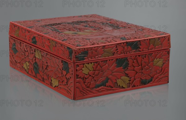 Box with a lid, Chinese art, Chinese culture, figural scenes, floral motifs, Qing 1644–1911, peonies, bamboos, geometric ornament, gourds, sages, scholars, fans, men. Decoration: on the lid a large reserve with a figural scene in a landscape; the rest of the background and sides decorated with floral motifs. Red, black, green, and yellow lacquer, carved. 19th century, China. Container. Height 13.0 cm, width 36.5 cm, depth 28.5 cm., A rectangular carved box sits on a reflective surface, shown from a low angled view. The box is predominantly red with carved floral patterns covering its top and sides, and a few carved leaves and petals are colored dark green and muted yellow. The carved surfaces have layered, sculpted textures with overlapping flower motifs and curved veining. The lid fits over the base with a narrow rim visible, and the reflection of the box appears faintly on the smooth surface beneath. The background is a uniform cool gray.
