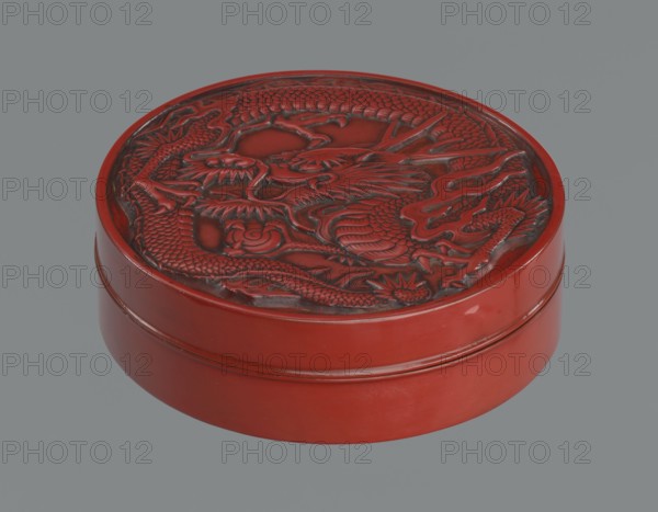 Box with a lid., Japanese, Meiji period 2nd half of the 19th century. Wooden core covered with lacquer; carved red lacquer exterior, black lacquer interior. Decoration: dragons, dragon with a pearl. Function: container. Dimensions: height 3.5 cm; diameter 10.9 cm; weight 149 g., A round lacquered box with a raised carved lid showing a coiled dragon motif, the box and carving predominantly red with darker red and black shading in the carved recesses; the exterior sides are smooth and glossy red; the background is a flat gray.