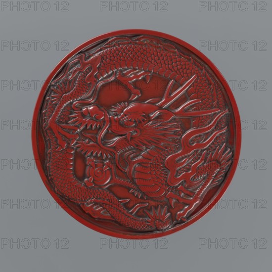 Box with a lid., Japanese; Meiji period 2nd half of the 19th century. Wooden core covered with lacquer; carved red lacquer exterior, black lacquer interior. Decoration: dragons, dragon with a pearl. Function: container. Height 3.5 cm; diameter 10.9 cm; weight 149 g., A circular carved object with a raised dragon motif occupying most of the surface, showing textured scales, a flowing mane, claws, and sinuous limbs; the carving has deep relief and glossy highlights. The object is predominantly red with darker red to near-black shadows in the recesses of the carving. The background behind the circular piece is a flat muted gray. The edges of the circular object are rounded and show the same red color with darker shading along the inner rim.