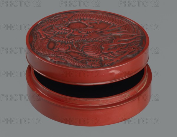 Box with a lid., Japanese, Meiji period 2nd half of the 19th century. Box with a wooden core covered with lacquer; carved red lacquer on the outside, black lacquer on the inside. Decoration: dragons dragon with a pearl. Function: container. Dimensions: height 3.5 cm; diameter 10.9 cm; weight 149 g., A round red lacquer box with a lid slightly ajar revealing a deep black interior, the lid featuring an embossed dragon design with textured scales and flowing mane details in darker red and black shading; the box surface shows glossy red highlights and subtle wear on edges, and the background is a flat gray.