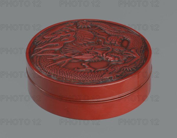 Box with a lid. Japanese, Meiji period 2nd half of the 19th century. Wooden core covered with lacquer; carved red lacquer on the outside, black lacquer on the inside. Decoration: dragons iconography, dragon with a pearl. Function: container. Height 3.5 cm; diameter 10.9 cm; weight 149 g., A round carved object with a lid featuring a raised dragon design, shown against a plain gray background; the object is predominantly red with darker red and black shading in the carved areas, and the sides have smooth glossy red surfaces.