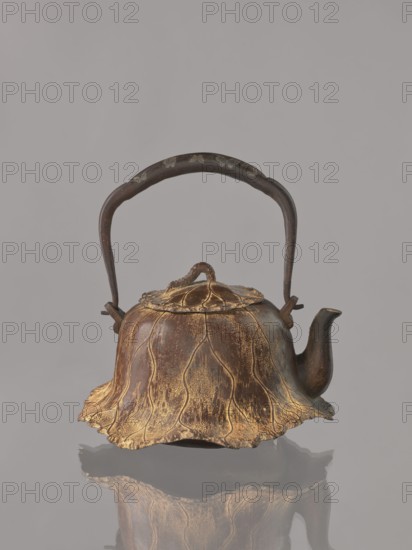 Teapot tetsubin with lid, lotuses and butterflies decoration, Japanese art, Meiji 1868–1912, tetsubin, 19th/20th century Meiji Period, Japan, vessel; height 20.0 cm, diameter 18.0 cm., A metal teapot with a dark brown handle and body that shows gold-colored highlights and textured, leaf-like ridges, a curved spout, a lid with a small looped knob, and an undulating, petal-shaped base edge; the colors visible include dark brown, gold, and gray, and the teapot sits on a reflective surface that mirrors its shape.