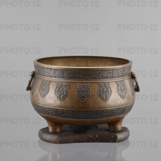 Three-legged incense burner, Tibetan, 19th century; craft; censers/ritual/ceremonial vessel; incense burner; height 25 cm, diameter 30.5 cm., A rounded metal vessel with a warm brown and bronze patina sits on a dark wooden base, showing a band of darker ornate raised patterns near its rim and another decorative band around its widest part, with a series of teardrop-shaped raised motifs around the midsection; the vessel has a smooth inner surface, a pair of ring handles attached at opposite sides, and three short rounded feet that rest on the wooden stand, with visible variations in sheen and subtle darker spots on the metal; the visible colors include brown, bronze, dark brown, and hints of black.