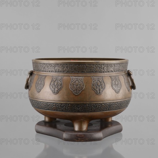 Three-legged incense burner, Tibetan, 19th century; craft metal censer/ritual/ceremonial vessel; incense burner; height 25 cm, diameter 30.5 cm;, A metal bowl with a rounded body and wide rim sits on a dark wooden stand; the bowl has a warm brown and bronze finish with darker engraved decorative bands around the top and lower midsection and a repeating teardrop-shaped motif around its sides, and a pair of ring handles attached to its upper sides; the wooden stand is dark brown with visible wood grain and a smooth, slightly curved shape; the background is a uniform light gray.