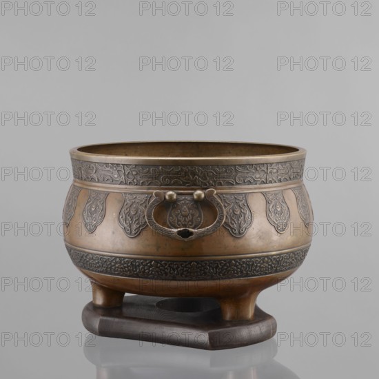 Three-legged incense burner, Tibetan, 19th century; craft; censers/ritual/ceremonial vessel; incense burner; height 25 cm, diameter 30.5 cm., The image shows a metallic bowl-like vessel with a rounded body and a wide open top, resting on a dark wooden stand; the vessel has ornate raised decorative bands and motifs around its upper and lower sections and a pair of circular handles with a ring between them on the front; visible colors include bronze and dark brown on the vessel and stand, with a neutral gray background.