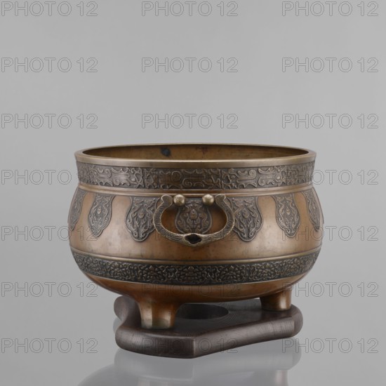 Three-legged incense burner, Tibetan, 19th century, craft; censers, ritual/ceremonial vessel; incense burner; height 25 cm, diameter 30.5 cm., A rounded metal vessel with a warm brown and darker brown patina sits on a dark brown carved wooden stand; the vessel has ornate raised decorative bands and repeating teardrop-shaped motifs around its body in darker brown, a raised rim with intricate patterned relief, a decorative ring handle on one side with two round knobs, and three short rounded feet supporting the bowl, all shown against a plain light gray background.