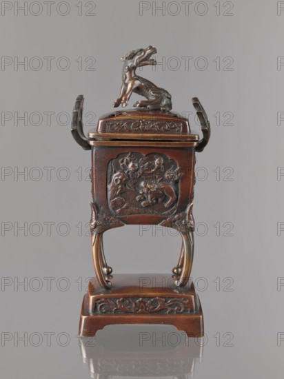 Incense burner koro with cover;, Incense burner in the shape of a cuboid box on four slightly arched legs standing on a tall, rectangular base. Panels with relief decoration of karashishi and peonies. Cover with four holes topped with a qilin. Bronze. Meiji period 2nd half of the 19th century, Japan. Height 22.0 cm; width 9.5 cm; depth 7.5 cm., A small ornate dark brown carved wooden container with a lid topped by a sculpted animal figure, shown against a neutral gray background; the container rests on an elevated base with curved legs and decorative panels featuring carved floral and animal motifs, and there are visible highlights of lighter brown and subtle darker brown patina across the surface.