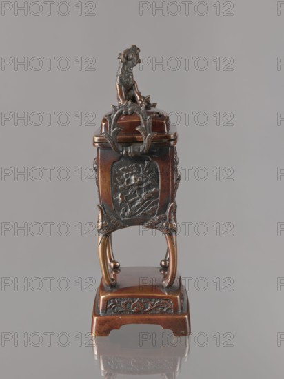 Incense burner koro with cover, Japanese, Meiji 1868–1912. Bronze vessel in the shape of a cuboid box on four slightly arched legs standing on a tall rectangular base. Panels with relief decoration of karashishi and peonies. Cover with four holes topped with a qilin. 2nd half of the 19th century. Height 22.0 cm; width 9.5 cm; depth 7.5 cm., A small ornate object stands against a plain gray background, primarily brown with darker bronze or blackened details; it has a carved animal figure on top in dark bronze tones, decorative curved metalwork in brown and dark bronze beneath it, and a relief panel on the front featuring a floral or botanical motif in dark bronze on a brown base. The piece rests on four curved legs that are brown with darker accents and sit on a rectangular brown platform that has darker carved ornamentation along its front edge. The surface shows polished brown wood tones and darker bronze highlights, and the object casts a faint reflection on the smooth surface below.