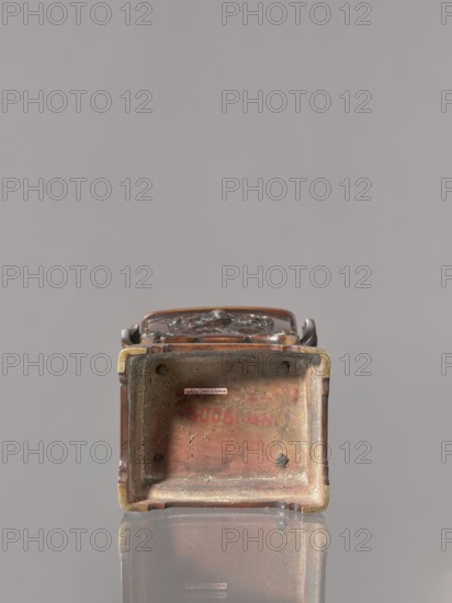 Incense burner kôro with cover, Japanese, Meiji 1868–1912. Incense burner in the shape of a cuboid box on four slightly arched legs standing on a tall, rectangular base. Decoration: panels with relief of karashishi and peonies. Cover with four holes topped with a qilin. Bronzes; peonies; flowers; karashishi iconography. 2nd half of the 19th century. Vessel. Height 22.0 cm; width 9.5 cm; depth 7.5 cm., A small rectangular metal object sits upright on a reflective surface, showing its open bottom interior; the object has a worn, mottled surface with shades of brown, copper, brass, and hints of greenish patina, and there are dark spots and rough texture inside. The exterior edges show darker brown and reddish tones with brass-colored corners, and a sculpted top edge with curved handles in darker metal is partially visible. The reflection below mirrors the colors and shapes, and the background is a smooth, neutral gray.