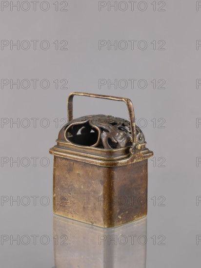 Hand warmer teaburi with lid, Japanese; Meiji period 1868–1912, 19th/20th century; bronze; rectangular vessel/container; decorated with landscapes and clouds; height 13.6 cm, width 10.9 cm, depth 8.2 cm., A small rectangular metal container with a hinged perforated lid and a thin arched handle sits on a reflective surface, showing a patina of brown and aged gold tones; the lid has ornate cut-out patterns and darker brown areas, while the body of the container displays worn brown and brassy gold patches with some darker spots and subtle surface imperfections, all set against a plain light gray background.