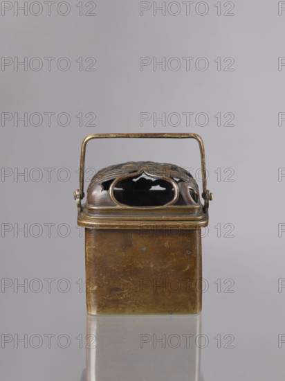 Hand warmer teaburi with lid, Japanese, Meiji period 1868–1912, 19th/20th century; rectangular bronze vessel/container decorated with landscapes and clouds; height 13.6 cm, width 10.9 cm, depth 8.2 cm., A small rectangular metal container with a hinged lid and a rectangular arched handle, shown against a plain light gray background; the container and handle are metallic with brownish-gold and darker bronze tones, showing areas of patina and wear, and the lid has an ornate cutout design with curved openings revealing a dark interior; the surface beneath the container is reflective, producing a soft mirrored image of the bottom edge.