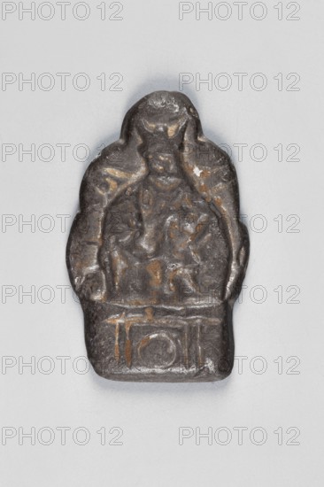 Thogcha with a representation of a human figure;, ; Tibetan Buddhist, Himalayan; human figures; 18th–19th century, Tibet; cult object; height 4.7 cm, width 3.0 cm, A small sculpted object sits against a light gray background, showing a worn figurative relief with a rounded top and a rectangular base. The surface of the object is primarily dark gray to almost black with patches of brownish and ochre wear that highlight shallow carved lines and shapes. The central portion suggests a seated figure within an arched outline, with raised areas that form a head and shoulders and lower raised areas that indicate a body and base. The edges are irregular and smoothed, and the overall texture appears rough and slightly pitted with some subtle reflective sheen on higher points. The visible colors are dark gray, black, brown, and ochre against the pale gray background.