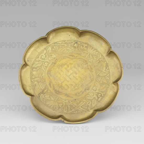 Platter, gift, plant ornament, happiness symbol, Chinese symbols, Korean culture, Korean art, hundred ancient baigu ornament, decoration: in the middle in a circle the fu sign happiness, symbols of prosperity and a plant ornament, early 20th century, Korea, vessel, height 15.0 cm, diameter 9.8 cm, A shallow metal dish with a scalloped rim shaped like a flower, shown against a neutral grey background; the dish has a golden yellow brass color with areas of darker patina and faint surface marks, and the interior base features an engraved circular decorative pattern of intertwining floral and geometric motifs in a slightly darker tone.