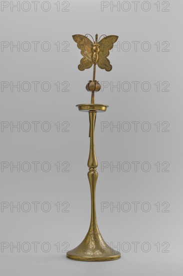 Candlestick, early 20th century, Korea; candlestick on a round base transitioning into a two-baluster shaft terminating in a flat, round sleeve with a smaller one in the shape of a multi-petaled flower bud placed in the center; decorated with butterflies and flowers; from a pair other no. SKAZsz 2509; height 39.7 cm., A tall, slender metallic object with a warm golden color stands against a plain light gray background; its flared base is decorated with etched swirling patterns, the narrow stem rising with smooth, rounded contours to a shallow dish-like platform, above which a thin rod supports a stylized flower bud and, at the top, a symmetrical butterfly-shaped ornament with delicate etched wing patterns and curled antennae, all sharing the same golden hue with subtle variations in sheen and small darker areas from patina.