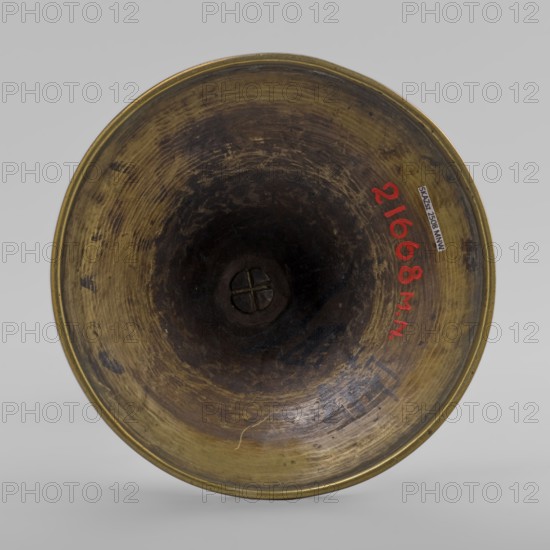 Candlestick, early 20th century, Korea; candlestick on a round base transitioning into a two-baluster shaft terminating in a flat, round sleeve with a smaller one in the shape of a multi-petaled flower bud placed in the center; decorated with butterflies and flowers; from a pair other number SKAZsz 2509; height 39.7 cm., A circular metallic object viewed from above with a concave interior surface showing worn textures and circular tool marks, the metal surface displaying shades of brass and brown with darker brown toward the center and some blackened areas, a small circular raised feature with a cross-like indentation at the center, a few spots of red paint on the interior rim, and a neutral light gray background.