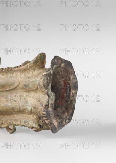 Guanyin; unknown creator; repossession Krakow; figurine; Guanyin iconography; Chinese art; Chinese culture; Chinese Buddhism; Ming 1368–1644; Buddhist art; goddess; bodhisattva iconography; mercy; natural patina; 16th century ; China; sculpture; height 39.7 cm, A close-up view of an aged, irregularly shaped metallic object lying on its side, showing a weathered surface with patina and pitting; the object displays tones of green, brown, gold, and dark gray with areas of lighter and darker discoloration; sculpted ridges and rounded protrusions run along its length, and one end has a hollowed, flared opening with rough, uneven edges; the background is a smooth gradient of light gray to white.