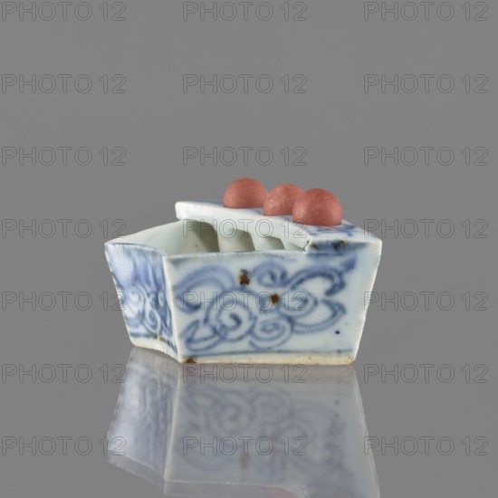 Vessel, ceramics, Chinese art, Chinese, plant ornaments, blue-and-white porcelain, brick-red balls fired from clay, early 20th century, China, vessel; height 3.1 cm, width 7.5 cm, depth 4.6 cm, A small ceramic object sits on a reflective surface against a gray background, shaped like a shallow rectangular boat with rounded inner dividers and a flat top that holds a group of three rounded beads. The ceramic has a white base color with blue painted swirling floral patterns on its sides and faint brown spots in places, and the beads on top are a muted reddish-pink. The surface reflects the object, mirroring the blue patterns and reddish-pink beads.