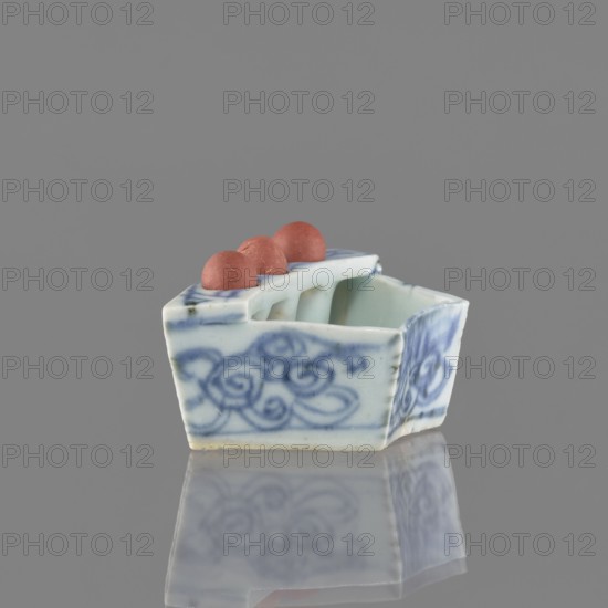Vessel, ceramics; Chinese blue-and-white porcelain with plant ornaments and brick-red fired clay balls; early 20th century, China; vessel; height 3.1 cm, width 7.5 cm, depth 4.6 cm., A small ceramic object with a glossy white surface and blue painted swirl patterns sits on a reflective surface, showing a faint mirrored image beneath. The object has an angular, boat-like shape with an open interior and a raised rim that contains a few rounded, matte red balls resting along the top. The visible colors are white, blue, red, and gray from the background and reflection.