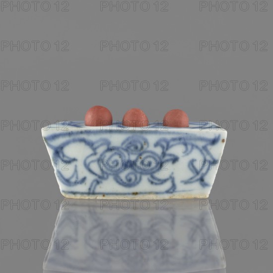 Vessel, ceramic, blue-and-white porcelain with plant ornaments and brick-red fired clay balls, early 20th century, China; height 3.1 cm, width 7.5 cm, depth 4.6 cm., A small rectangular ceramic object with sloping sides sits centered against a solid grey background, decorated with blue swirling floral-like brushwork on a white base; three round reddish-brown spheres rest in shallow depressions along the top edge, and the object and its decorative pattern are reflected faintly on the glossy surface beneath.
