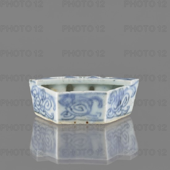 Vessel, gift, repossession Krakow, ceramics, Chinese art, Chinese culture, plant ornaments, blue-and-white porcelain, brick-red balls fired from clay, early 20th century, China, vessel, height 2.6 cm, width 7.4 cm, depth 4.7 cm, A small hexagonal ceramic dish with scalloped top edges, glazed in pale white and decorated with blue painted swirling floral motifs on its sides, sitting on a reflective surface that mirrors the dish; visible colors include white, blue, gray, and a few small brown spots on the base.