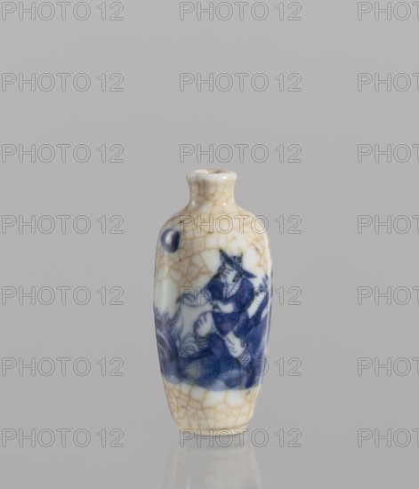 Flask-style snuffbox of elongated ovoid shape with a narrow short neck. Blue-and-white porcelain with bluish glaze and cracklework highlighted in brown paint, depicting figural scenes. 19th century, China. Height 7.3 cm; diameter 3.5 cm., A small ceramic bottle with a narrow neck and rounded shoulders stands against a plain gray background, its surface showing a network of fine crackled lines in tan and beige over a white base. A band of deep blue painted decoration wraps around the midsection, depicting a stylized human figure wearing a hat and flowing garments amid abstract shapes and curved motifs. The bottle’s foot and shoulder areas display the same crackled tan pattern, and the overall colors visible are white, tan, beige, and deep blue.