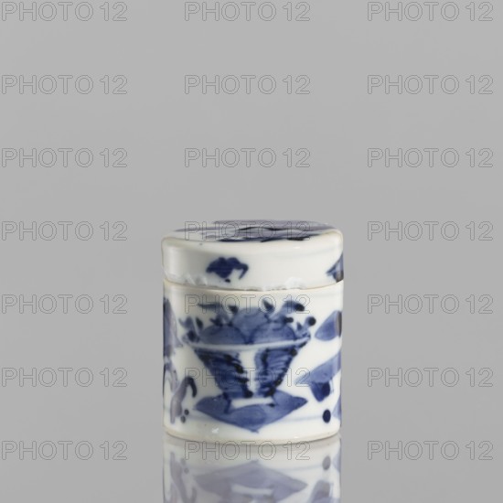 Cylindrical jar with a lid; ceramics, blue-and-white porcelain; cylindrical vessel with attached lid; thick porcelain body with bluish-tinted paste; decoration painted underglaze with cobalt, thickly applied in places; glaze with a grayish hue; ornament motif: baigu one hundred antiquities; Chinese; 19th century; height 5 cm, diameter 4.5 cm., A small cylindrical ceramic container with a fitted lid sits against a plain light gray background, showing a glossy white surface decorated with hand-painted blue motifs; the lid and body display organic, brushstroke patterns including floral and leaf-like shapes, with several areas of darker and lighter blue creating a painterly effect, and the object casts a faint reflection on the smooth surface beneath it.