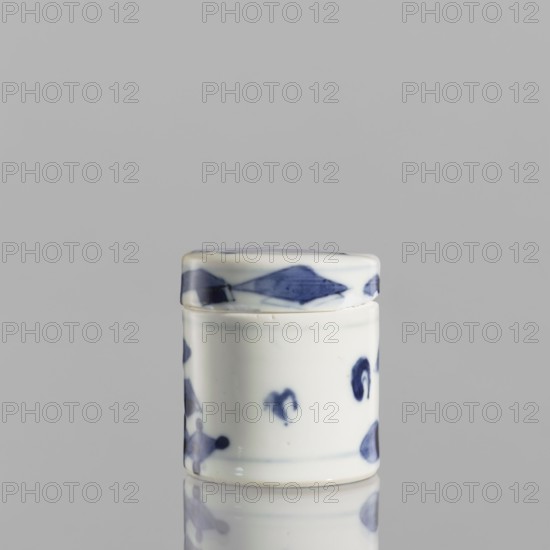 Cylindrical jar with a lid; ceramics; blue-and-white porcelain; cylindrical vessel with attached lid; thick porcelain body with bluish-tinted paste; decoration painted underglaze with cobalt, thickly applied in places; glaze with a grayish hue; style: Chinese; 19th century; height 5 cm, diameter 4.5 cm., A small cylindrical ceramic container with a fitted lid sits on a reflective surface. The container is primarily white with several blue painted decorative shapes and smudges around its sides and on the lid, including a band of blue shapes near the top edge. The glaze gives the surface a glossy finish, and the reflection of the container appears faintly on the surface below. The background is a flat neutral gray.