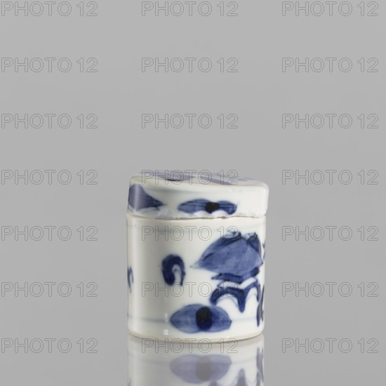 Cylindrical jar with a lid; ceramics; blue-and-white porcelain; Chinese, 19th century. Thick porcelain body with bluish-tinted paste; decoration painted underglaze with cobalt, thickly applied in places; glaze with a grayish hue. Cylindrical vessel with attached lid; height 5 cm, diameter 4.5 cm. Contains motif one hundred antiquities baigu., A small cylindrical ceramic container with a fitted lid, both surfaces glazed in white with hand-painted blue floral and abstract brushwork; the container and lid show rounded edges and a glossy finish, and the object sits on a reflective surface that mirrors its blue and white tones against a plain light gray background.