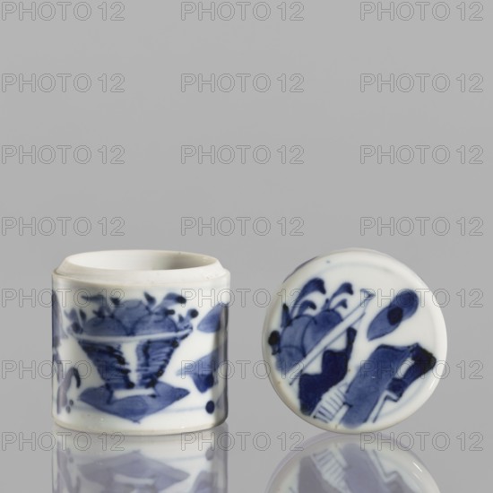 Cylindrical jar with a lid; ceramics; blue-and-white porcelain; Chinese; 19th century. Cylindrical vessel with attached lid; thick porcelain body with bluish-tinted paste; decoration painted underglaze with cobalt, thickly applied in places; glaze with a grayish hue. Height 5 cm; diameter 4.5 cm., A small cylindrical ceramic container with a separate round lid sits on a reflective surface against a plain light gray background. The container and lid are white with hand-painted blue designs; the container shows a stylized floral or leafy motif wrapping around its side, and the lid displays abstract organic shapes and brushstrokes in varying shades of blue. The ceramic has a glossy finish, and both pieces cast soft reflections on the surface below. The visible colors are white, several shades of blue, light gray, and subtle reflections of those colors.