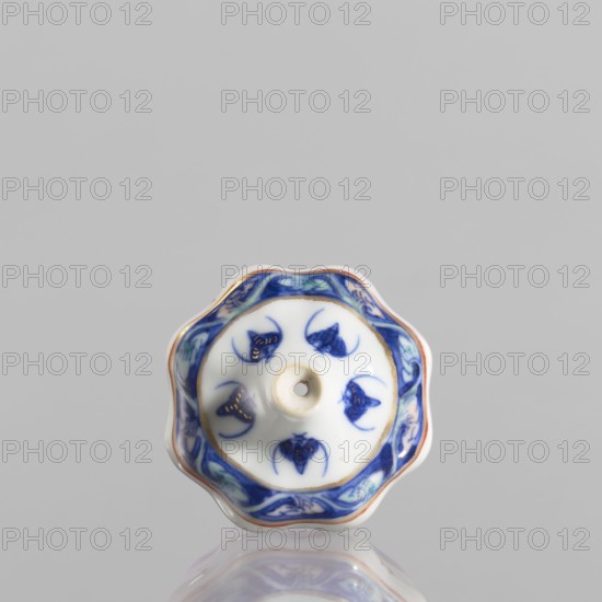 Spice vessel, 19th century China, blue-and-white porcelain; vessel no stopper; height 4.65 cm, diameter 5.5 cm; decorated with bats; used for spices., A small scalloped ceramic object with a glossy white base and a central raised hole, decorated with a blue border featuring floral-like motifs and several blue stylized shapes on the white surface, with touches of gold accenting parts of the design and a thin reddish-brown rim, all reflected faintly on the glossy surface beneath; visible colors include white, various blues, gold, reddish-brown, and a hint of green.