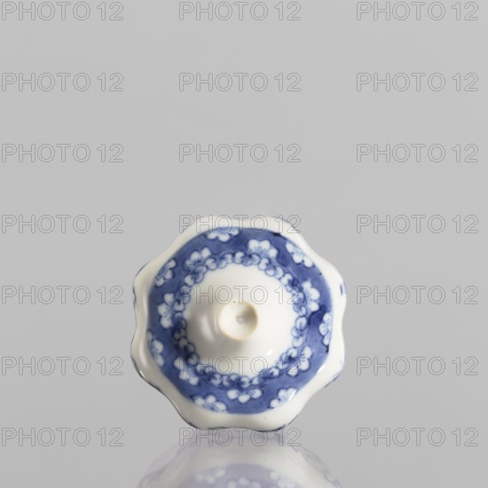 Small spice vessel, blue-and-white porcelain, no stopper, 19th century, China; vessel height 4.6 cm, diameter 5.5 cm., A small ceramic object viewed from above with a scalloped rim and a central raised circular depression, predominantly white with a blue floral and chain-like decorative band around the center; the background is a flat light gray and the object casts a soft reflection on the glossy surface below, showing colors of white, blue, light gray, and a touch of pale beige in the depression.