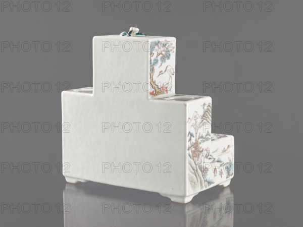 Pedestal for a writing set three-stage, ceramics with colored porcelain in Qing style late 19th century, China. Decorated with flowers, figural scenes, butterflies, and landscape motifs; raised central part. Function: writing accessories. Dimensions: height 18.9 cm, width 17.9 cm, depth 11.0 cm., A white stepped ceramic object with a glossy finish sits on a reflective surface against a gray background; the visible faces feature delicate painted scenes in muted colors including blue, green, brown, pink, and red, showing natural elements like trees, mountains, and birds, and small human figures in traditional clothing; the top has a small sculpted decorative element in green and white, and the object rests on short feet with slight shadows and reflections beneath.