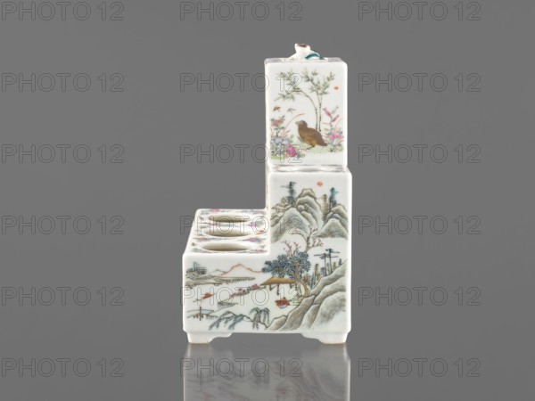 Pedestal for a writing set three-stage, ceramics, colored porcelain; decorated with flowers, figural scenes, butterflies, and landscape motifs in Qing style Qing, 1644–1911, raised central part; late 19th century, China; writing accessory; height 18.9 cm, width 17.9 cm, depth 11.0 cm., A small L-shaped ceramic object with a glossy white surface, decorated with painted scenes in colors including brown, green, pink, blue, orange, and black; the taller vertical section shows a bird among foliage and flowers with green leaves and pink blossoms, while the lower horizontal section features a landscape of mountains, trees, and a small pavilion rendered in muted greens, blues, and browns with touches of pink and orange; the top of the horizontal section has a row of round openings rimmed by floral-painted edges, and the piece rests on short feet with its reflection faintly visible on a smooth gray surface.