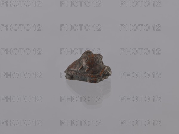 Group of turtles; okimono; creator, ceramic figurine; Meiji period 1868–1912, 19th/20th century, Japan; Japanese art; height 2.8 cm, width 3.5 cm, depth 5.5 cm., A small carved object resembling a turtle sits centered on a smooth reflective surface, showing a faint mirrored reflection beneath it; the object displays dark brown and reddish-brown tones with some blackened areas and visible carved lines and textured details on its shell and body, while the background and surface are a uniform light gray.