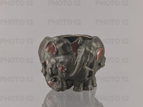 Flowerpot, gift, Japanese art, Meiji period late 19th century, 1868–1912, Japan; bronze flowerpot/jardiniere/vessel with animal motifs elephants, copper-plated vessel parts; height 16.0 cm, width 23.0 cm., A sculptural object shaped like a group of elephants forming a rounded vessel, with textured dark bronze-gray surfaces and reddish-brown inner ear areas, sitting on a reflective surface that mirrors its underside; the background is a smooth light gray.