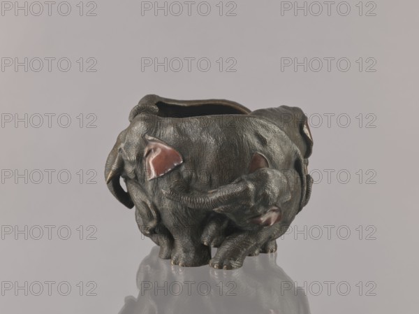 Flowerpot, Genius, Mieczyslaw 1853–1920, Japanese art, Meiji period late 19th century, 1868–1912, Japan; bronze and copper-plated vessel parts; jardinière/vessel with animals elephants; height 16.0 cm, width 23.0 cm., A dark sculptural object shaped like a group of elephants arranged in a circular formation is visible against a pale gray background; the sculpture has a textured dark brown to black surface with glossy reddish-brown accents on several ear-like shapes, a smooth interior opening at the top, curved trunks and rounded legs, and a reflective surface beneath that produces a faint mirrored reflection.