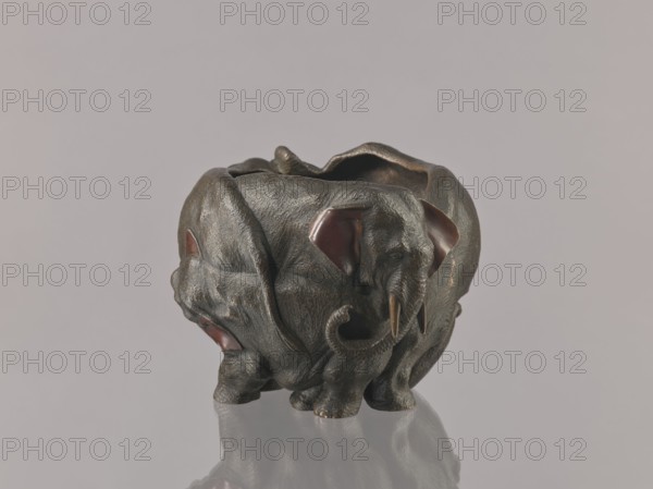 Flowerpot, gift, Japanese art, Meiji 1868–1912, bronzes, flowerpot/jardiniere/vase with animal elephant motif, copper-plated vessel parts, late 19th century Meiji period, Japan; vessel; height 16.0 cm, width 23.0 cm, A sculpted object shaped like an elephant with textured surfaces and a rounded hollow top, rendered in dark bronze tones with areas of brown and subtle gold highlights, showing reddish-brown inner ear details and darker shadowed recesses, positioned on a reflective surface with pale gray and muted beige background colors.
