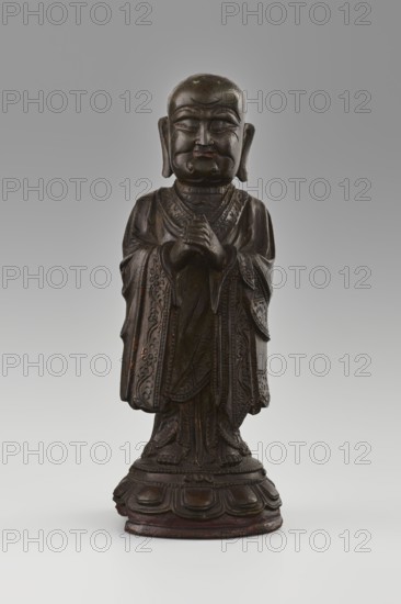 Monk, ; Chinese; Qing 1644–1911; 19th/20th century. Sculpture of a monk/arhat Luohan in Buddhist art iconography, seated on a round low lotus pedestal with lotuses. Terracotta/other material unspecified. Height 24.7 cm; diameter 9 cm., A dark brown carved wooden figure stands on a rounded base with layered petal-like shapes, wearing a long robe with detailed carved patterns and flowing sleeves, hands held together at chest level, a rounded bald head with pronounced facial features including closed eyes and a slight smile, elongated earlobes, and subtle wear showing lighter brown highlights and traces of reddish tones against a neutral gray background.