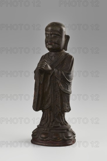 Monk, Papal, Chinese art; Chinese culture, Qing 1644–1911; figurine of a Buddhist monk/arhat Luohan seated on a round lotus pedestal lotus throne; sculpture, 19th/20th century, China; height 24.7 cm, diameter 9 cm., A dark brown carved wooden standing figure wearing layered draped robes with ornate carved patterns, shown against a smooth gray background; the figure has a rounded bald head, closed eyes, pronounced facial features, and hands held together in front; the carved base beneath the figure has a petal-like design and shows hints of reddish-brown and black tones.