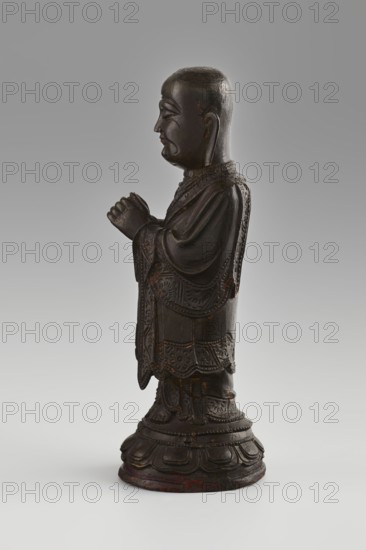 Monk, unknown role classification of creator, craftsman author; Papal, Leon Ildefons 1859-1917; revindication Krakow; Chinese art; Chinese culture; Qing 1644-1911; figurines; Buddhist art; Chinese Buddhism; monks; arhat iconography; Luohan iconography; lotus throne iconography; lotuses; Round lotus pedestal, low.; 19th/20th century; China place of origin; sculpture; height 24.7 cm; dia. 9 cm, A dark brown carved wooden standing figure shown in profile on a rounded ornate base, wearing layered robes with carved decorative patterns, hands held together near the chest, a smooth rounded head with carved facial features and closed eyes, small ears, and visible wear with lighter brown and reddish spots; the background is a gradient of gray and the surface beneath is white-gray.