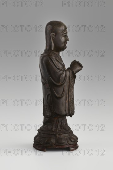 Monk, unknown role classification of creator, craftsman author; revindication Krakow - collection removed. Chinese art; Chinese culture; Qing 1644–1911; figurine; Buddhist art; Chinese Buddhism; monk; arhat Luohan iconography; lotus throne; lotuses; round low lotus pedestal. 19th/20th century, China. Sculpture; height 24.7 cm; diameter 9 cm., A carved dark brown wooden statue of a standing robed figure shown in profile facing right, with a smooth rounded head, elongated ears, closed eyes, and a serene facial expression, the hands held together near the chest, the flowing robes detailed with carved folds and patterned borders, the figure standing on a carved rounded base with layered petal-like motifs; visible colors include dark brown, with small areas of reddish-brown and faint traces of lighter brown on the surface, set against a neutral pale gray background.