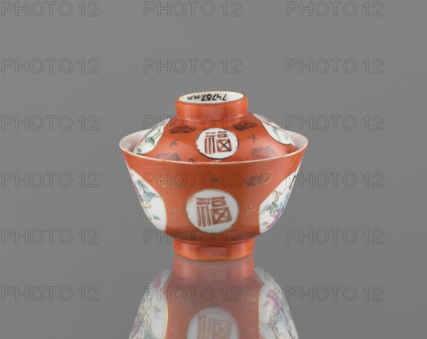 Bowl with a lid, Qing dynasty Tongzhi period, 1862–1874, Chinese, colored porcelain, ceramics; decorated with bats and swastikas and genre scenes, inscription/decoration note: In the background of the decoration loosely placed bats and swastikas. Vessel; height 8.6 cm, diameter 10.3 cm., A small lidded ceramic bowl with a rounded foot and flared rim sits centered on a reflective surface, showing a mirrored image below. The bowl is primarily a warm orange color with white circular panels around its body and lid that contain decorative motifs. There are accents of gold outlining small cloud-like shapes and other tiny decorative marks on the orange background. Within the white panels are painted scenes and figures in colors including green, pink, purple, and pale blue, with some black or dark brown fine lines for detailing. The interior visible at the rim is white, and the overall background behind the object is a flat gray.