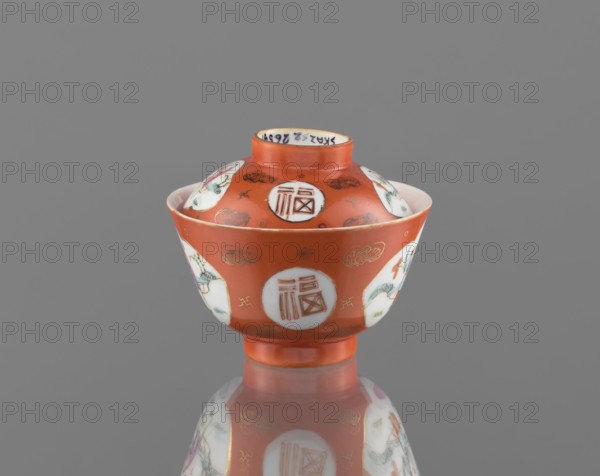 Bowl with a lid, ; Qing dynasty, Tongzhi period 1862–1874, China; colored porcelain; vessel with decoration of loosely placed bats and swastikas in the background and genre scenes; height 8.6 cm, diameter 10.3 cm., A small bowl with a lid sits centered against a neutral gray background, the bowl and lid predominantly a warm reddish-orange with glossy finish and a reflective surface beneath showing a faint mirror image. The rim, interior of some circular medallions, and portions of decorative panels are white, and the decoration includes painted motifs in green, pale blue, pink, and gold. The bowl has a short foot and a rounded body that tapers slightly inward toward the rim, and the lid has a small raised central knob area. Several circular white medallions with contained linear designs and a few oval panels with floral or figurative scenes encircle the exterior, and fine gold accents and small cloudlike gold motifs are scattered across the reddish-orange ground. The overall compositio...