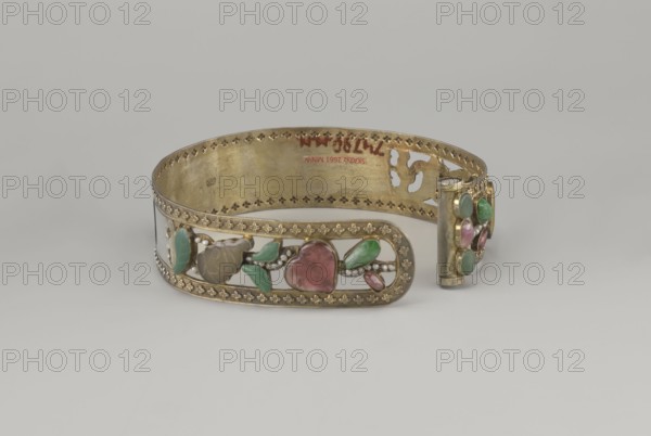 Headband, ; plant ornament; Chinese; Chinese art; Qing 1644–1911; head ornament; openwork inlaid with glass, semi-precious stones, river pearls and beads; 19th century; China; height 3.3 cm; diameter 12 cm., A metal bracelet with a gold tone band is shown resting on a light gray surface against a neutral gray background; the band has decorative perforated edging and an openwork panel featuring carved and polished stones in green, pink, and brown hues set among small bead-like details, with additional smooth green and pink stones on a cylindrical clasp element, and the inner surface of the band shows variations in the gold tone finish.
